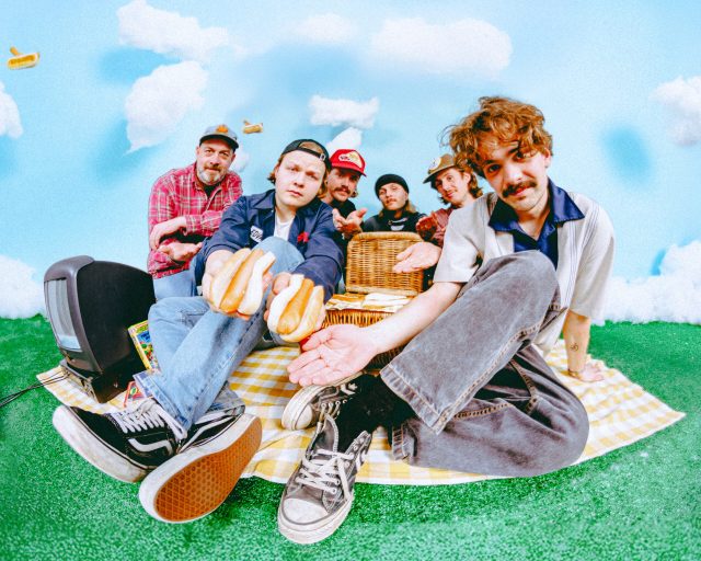 Her ser vi seks gutter som sitter på et piknikpledd. De deler pølser i brød.