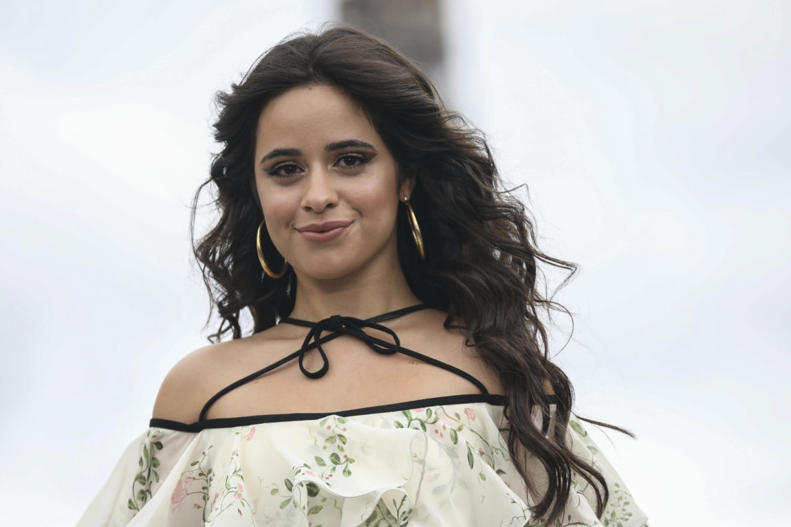 Camila Cabello har på seg to store gull øredobber. Hun har også en hvit topp med grønne blomster på. Hun smiler lurt inn i kameraet.