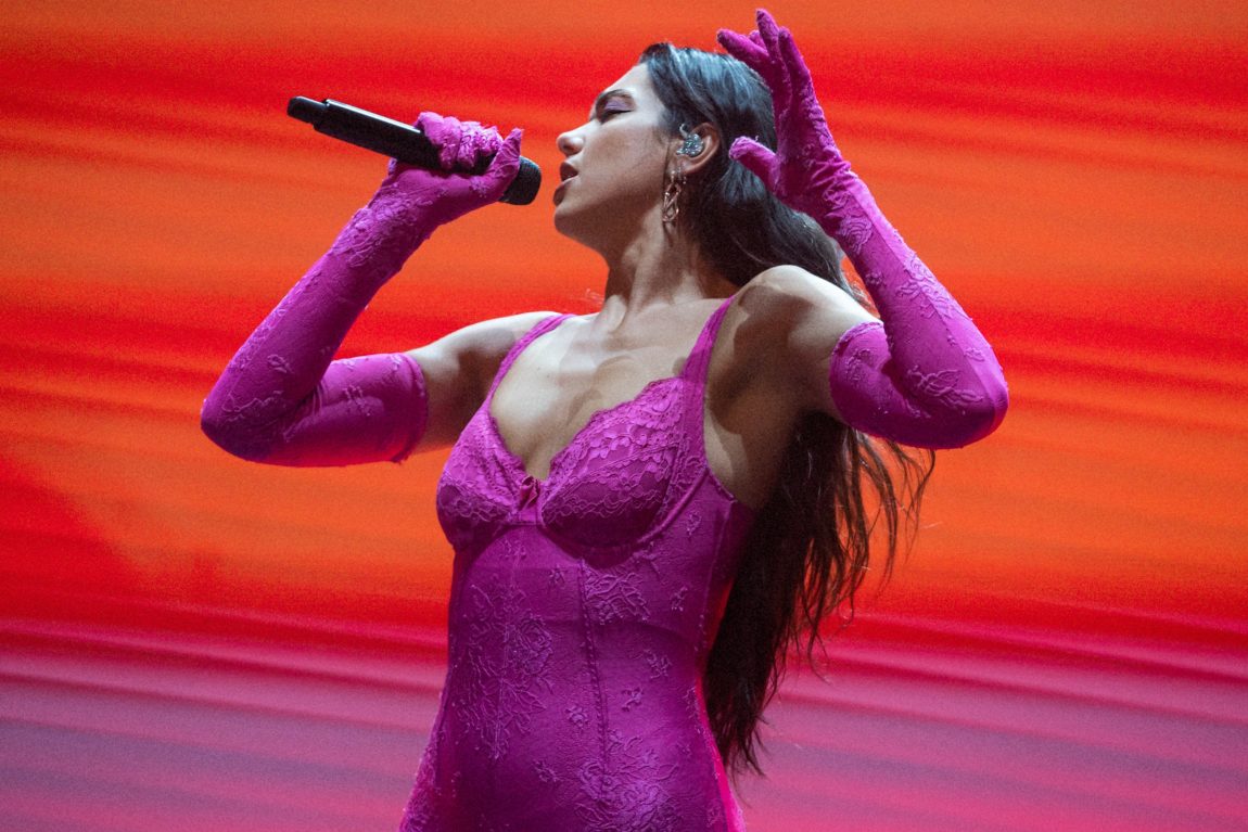 Dua Lipa i rosa kjole står foran en oransje og lille vegg. Hun holder en mikrofon tett inntill munnen.