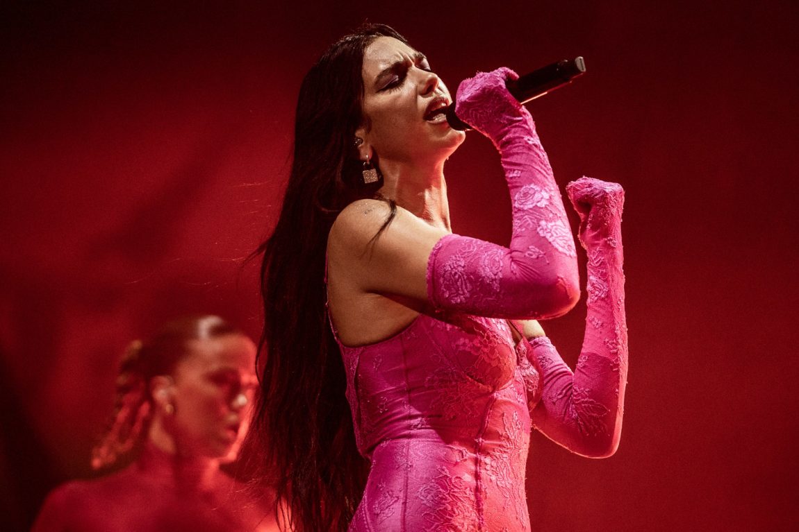 Dua Lipa synger ektefølt foran en rød vegg. I den rosa kjolen. I bakgrunnen er det en dame som ser skremt ut.