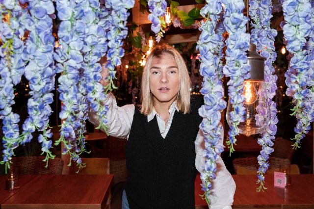 Ola står bak hengende lilla blomster inne på en restaurant. Han er ikledd en hvit skjorte og en sort strikkevest. Han ser med alvorlige øyne rett i kamera, ansiktet er pent sminket og øyenbrynene veldig symmetriske. Han har blondt hår til skuldrene.