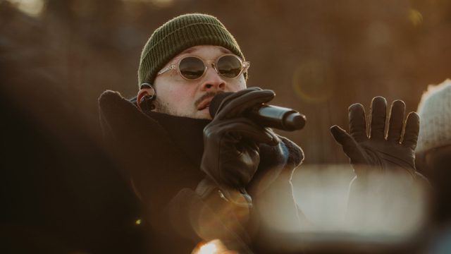 Stig Brenner holder mikrofonen tett til munnen. Han har på seg sorte skinnhansker, blikket er rettet fremover under et par runde solbriller. På hodet har han en mørkegrønn lue som sitter over ørene. En stor, sort krage omkranser hodet hans.