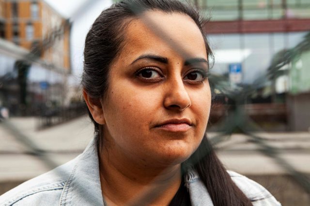 Kiran Anwar ser gjennom et stålgjerde, hun er alvorlig i blikket og ser rett i kameraet. Hun har dongerijakke på.