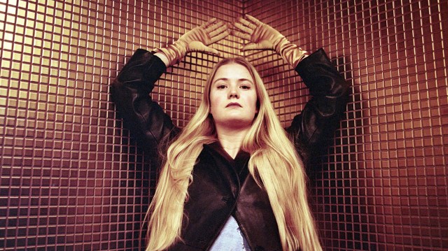 Artisten Amalie Holt Kleive står i et rom med små brune fliser og folder hendene sine over hodet, mens hun ser inn i kamera