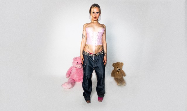 Sa_g har på seg baggie bukser og korsett. Hun står i et hvitt rom med teddy bjørner på hver side.