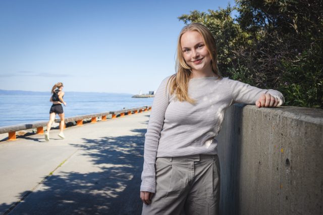 Blond jente står ved murvegg. Jogger langs strandprommenade i bakgrunn. Blå himmel og sol ved sjøen.