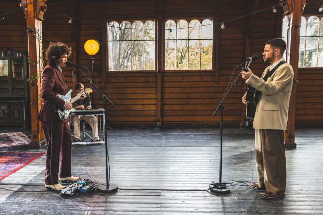 Fay Wildhagen og Chris Holsten står mot hverandre med hver sin mikrofon og gitar. Fay har på seg en lilla dress. Chris har på seg en beige dressjakke og brune bukser.