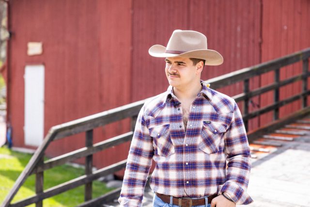 Tobias Sten har på seg en rutete skjorte, cowboyhatt og skjorta ned i buksa. Han går nedover en bondegård.