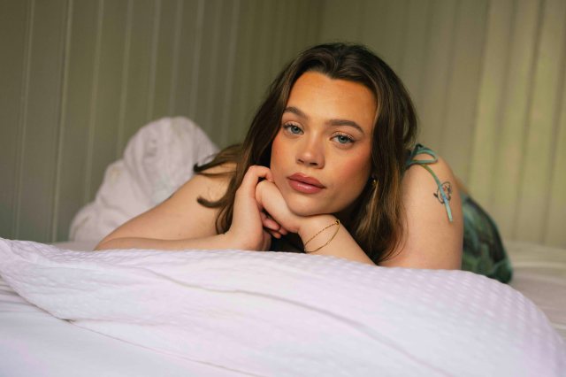 Emma Snoen ligger i senga og ser sårbart inn i kamera. Hun har brundt hår som henger ned på sidene.