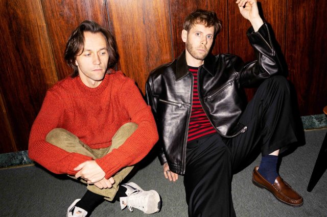 Sondre Lerche og Lars Vaular sitter på bakken og ser betenkte ut. Sondre har på seg en rød ullgenser og en grønn bukse. Lars har på seg en svart skinnjakke og en stripete rød og svart genser.