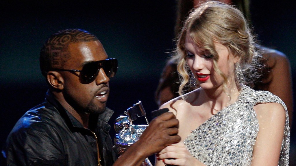 Et bilde av Kanye West som griper mikrofonen ut av hendene på Taylor Swift. Kanye har på seg solbriller og sort jakke. Taylor har på seg en glitrende, sølvarget kjole.