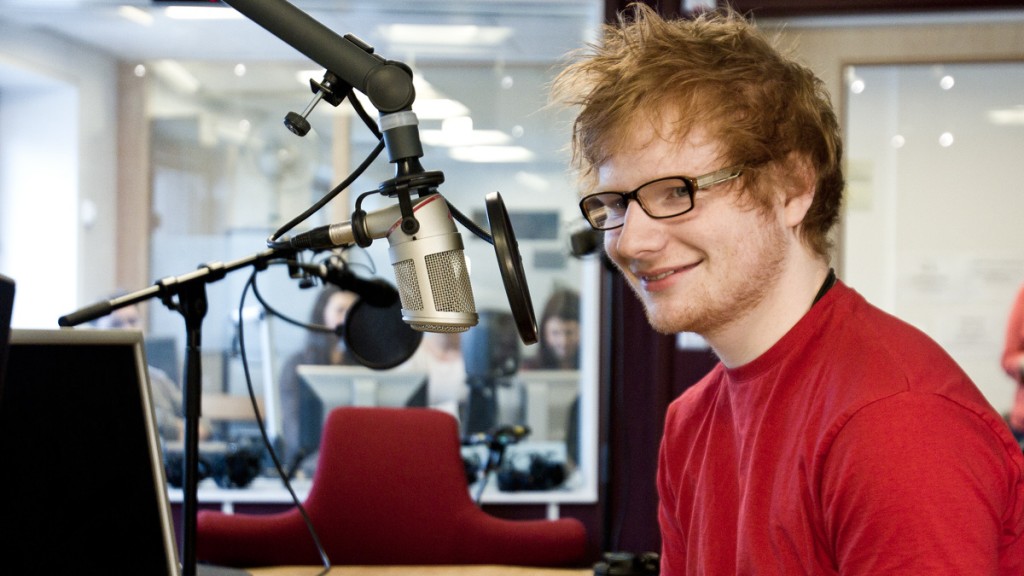 Et godt gammelt bilde av Ed Sheeran. Han har på seg en rød t-skjorte som matcher håret. Han har også briller og snakker inn i en radiomikrofon. 