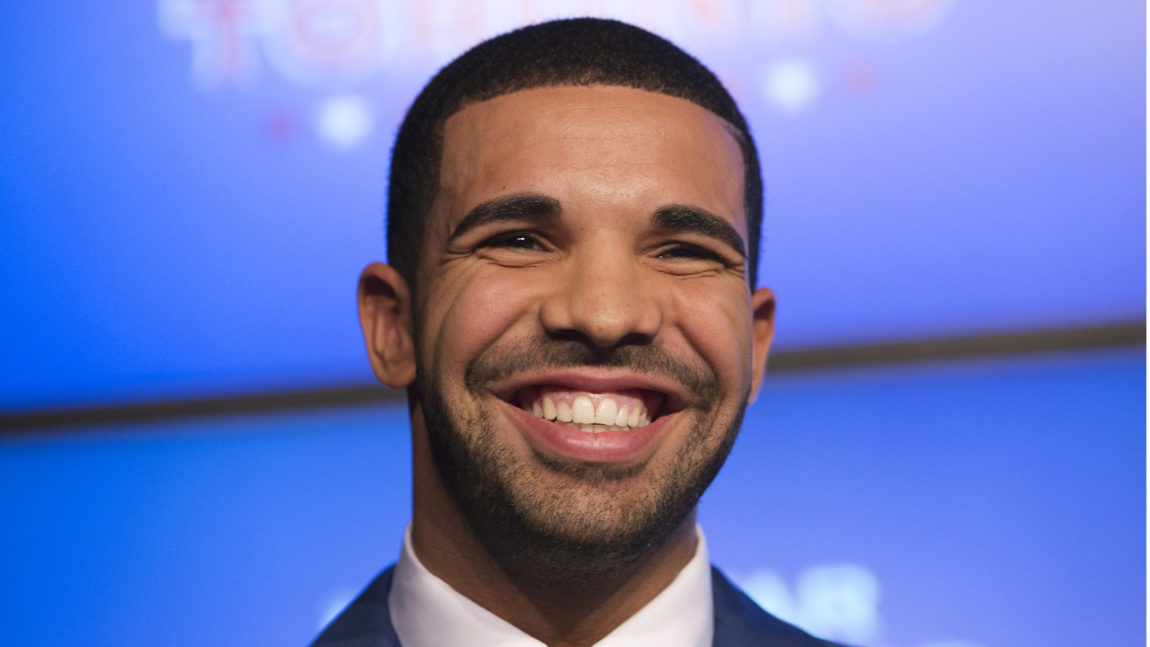 Drake gliser og viser tennene. Han har på seg en dress og står foran en blå skjerm.