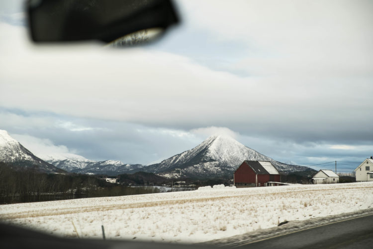 Utsikt fra forsetet i en bil. Til venstre er det to fjell med snø på toppen. Himmelen er både blå og grå av skyer. Til høyre skimtes en gård, med en rød låve og to hvite hus. Mellom gården og veien er et stort snødekt jorde.