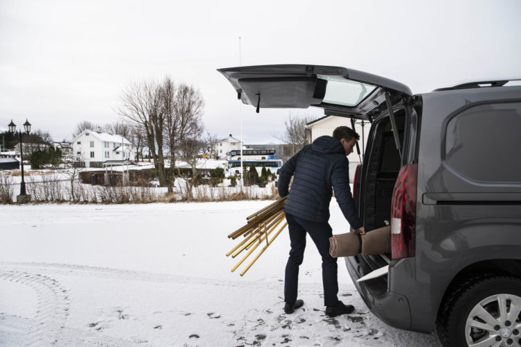 Til venstre er en parkeringsfplass med et tynt lag med snø, mens nabolag av hvite hus og to hvite og blå busser skimtes i bakgrunnen sammen med noen trær. Til høyre står Kristians grå bil, med bagasjerommet åpent. Kristian, iført blå boblejakke, drar ut en brun pakke med den høyre hånde og holder gullstativer i den venstre.