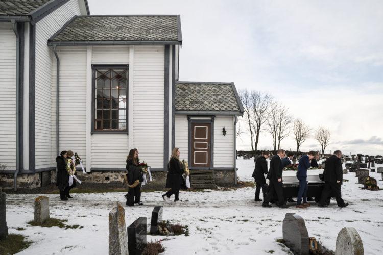 Til venstre er en hvit kirke med lyseblå kanter. Begravelsesfølge kledd i sort går nedover kirkegården med blomster. Til venstre bærer seks menn en hvit kiste.