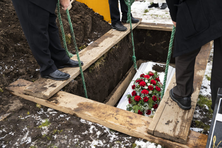 Tre menn legger kista i graven med grønne tau. De har alle svarte dressbukser og sko. På toppen av den hvite kista ligger en stor bukett med røde roser.