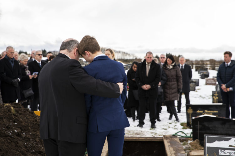 Til venstre står en eldre mann med ryggen til, ikledd sort blazer. Han holder rundt en yngre gutt med den høyre armen, mens de ser ned på graven og sørger. Gutten til høyre er ikledd blå dress.