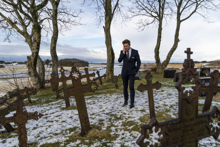 Kristian står til høyre, på baksiden av kirkegården, iført mørkeblå dress. Han snakker i mobiltelefonen og holder den med høyre hånd, mens han ser ned i grønt gress fylt av snø. Rundt han er gravstøtter av metallkors. I bakgrunnen er det åker og hav.