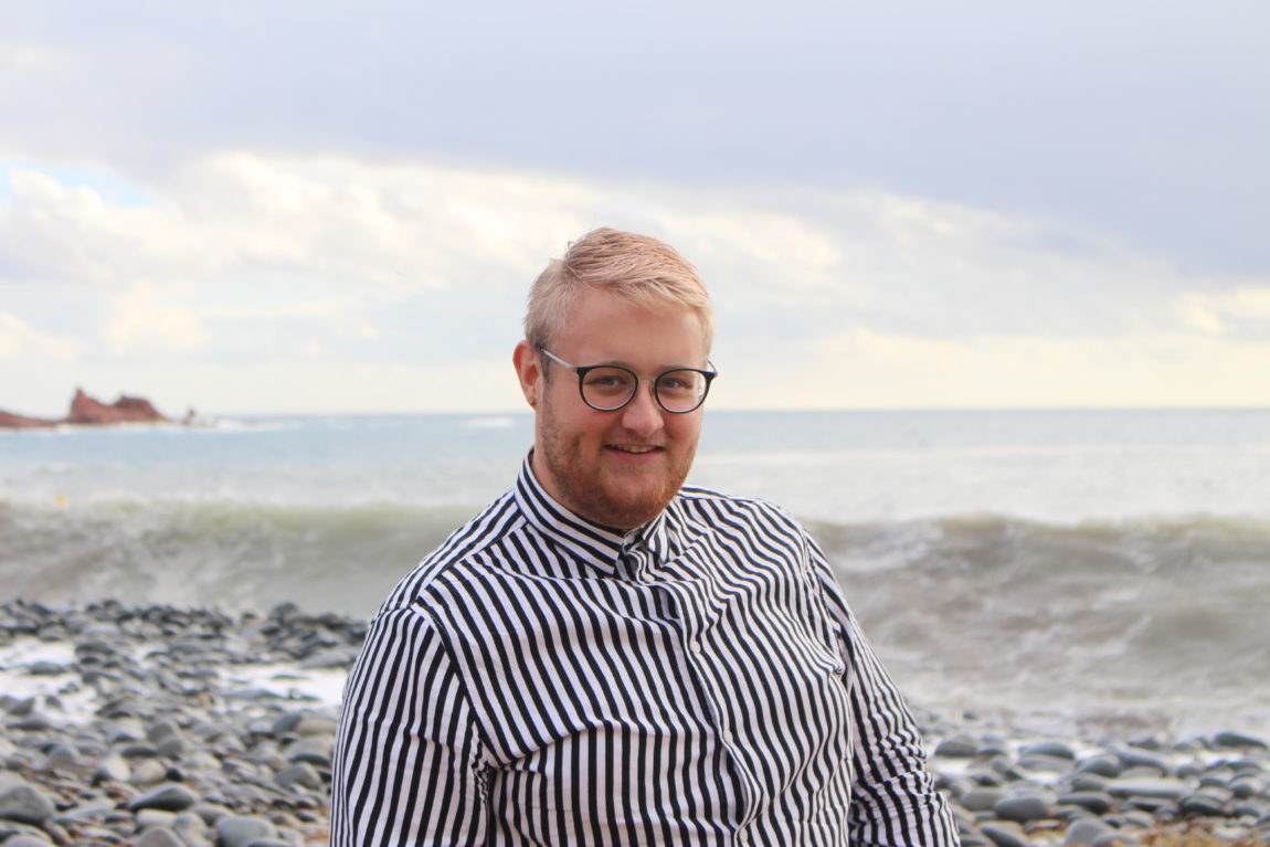 Ole står på en steinstrand med hav og bølger bak seg. Han smiler til kameraet og ha på seg en stripete hvit og mørkeblå skjorte. 