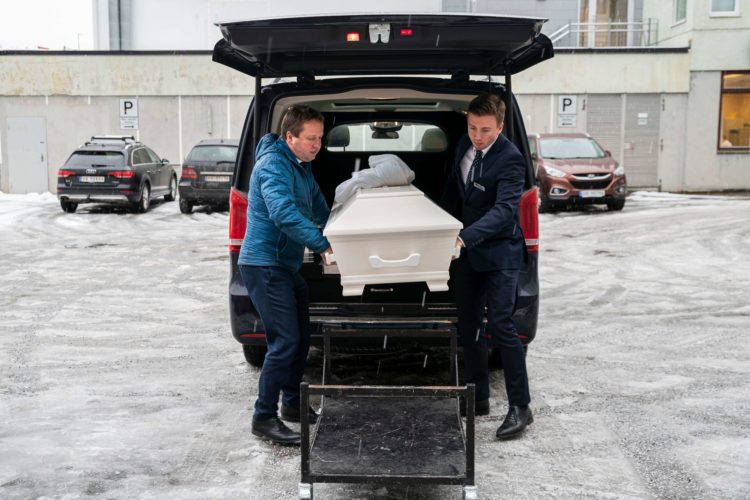 Faren til kristian og Kristian bærer ut en kiste fra bagasjerommet. Far til venstre er ikledd lyseblå jakke, sønn til høyre,  iført mørkelbå dress. På bakken er det lett snø og bak dem en parkeringsplass med biler.