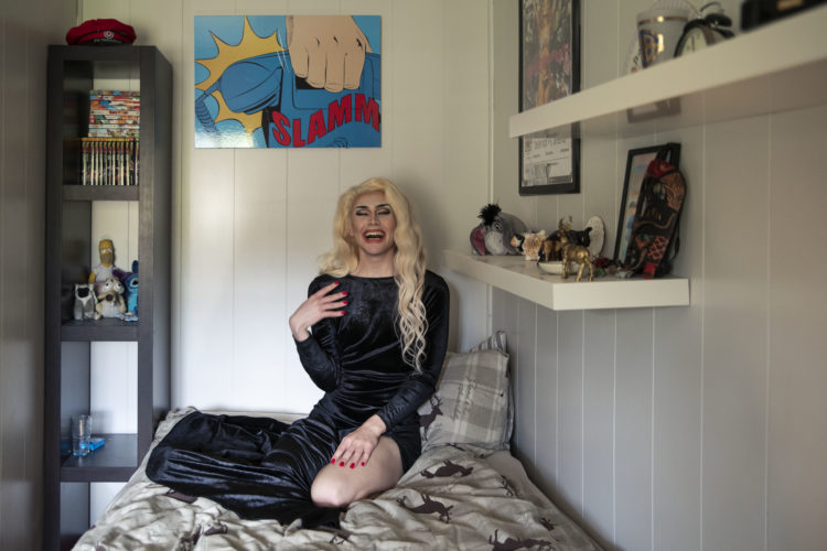 Sondre sitter på senga lent inntill veggen med det blå bildet. Han smiler bredt, er iført sort kjole og blond parykk og sminke. Til venstre er en sort hylle med tegneseriepockets og disne-kosedyr.