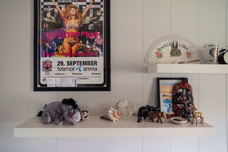 Hvit vegg med hvite hyller på Sondres rom. Til venstre henger en plakat av Lady Gaga hvor hun hever armene i været og smiler. Det er en reklameplakat for konserten hennes. På hylla under ligger en Tussi-bamse, strandskjell, treelefanter og en afrikansk maske. På hylla over ligger en håndvifte, et krus og en gammeldags vekkerklokke.