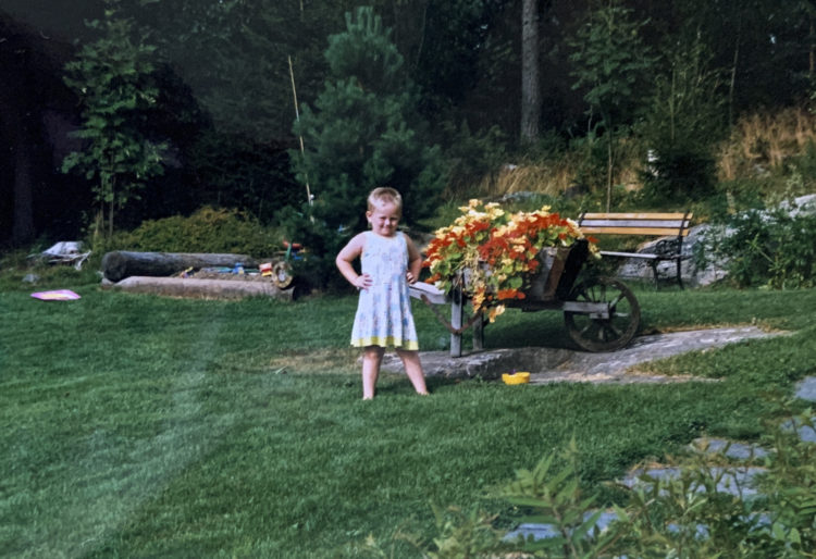 Barndomsbilde tatt med analogt kamera. Sondre står midt i bildet ved siden av gule og røde blomster. Han har hendene i sida, og smiler. På seg er en grønn-blå kjole med gule og hvite blomster. Alt rundt han er grønt.