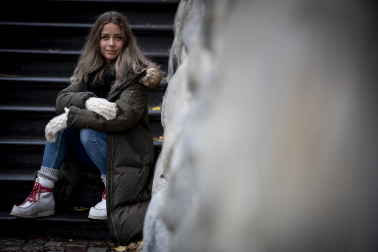 Natalie sitter til venstre i bildet på en steintrapp. Hun ser rett i kamera, med et lurt smil. Det lange brune håret er utslått, og hun er godt kledd for en kald høstdag i Umeå. 