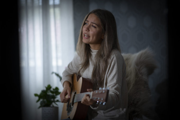 Natalie sitter hjemme i stuen sin i Umeå. Hun holder og spiller en gitar, og ser ut til å synge. 