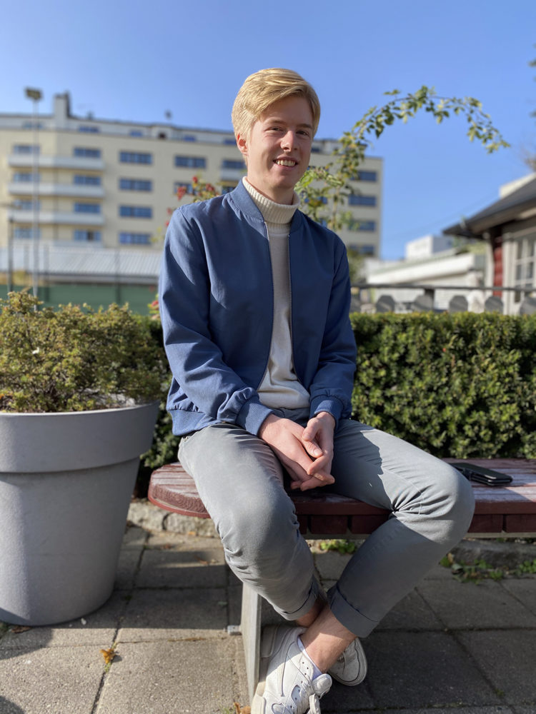 Eit bilete av ein ung student. Han smiler. Han har blondt hår. Han har på seg kvit genser og blå jakke.