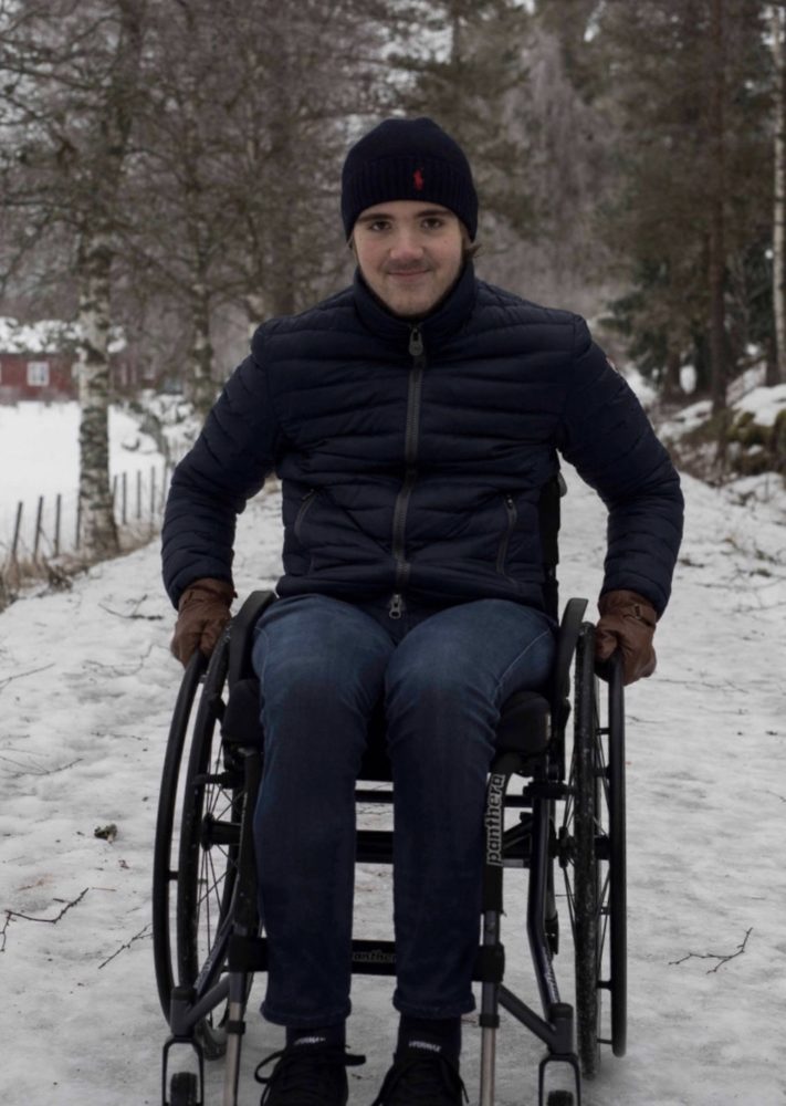 Marcus smiler til kamera. Han held fast i hjula på rullestolen. Det er eit tynt lag med snø på bakken rundt han. 