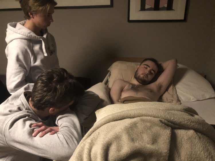 Marcus ligg med augo att på ei seng i stova. Han har armen bak hovudet, og det ser ut som om han søv. To kompisar våkar over han, Torjus ligg med armane på sengekanten og ser på vennen sin. 