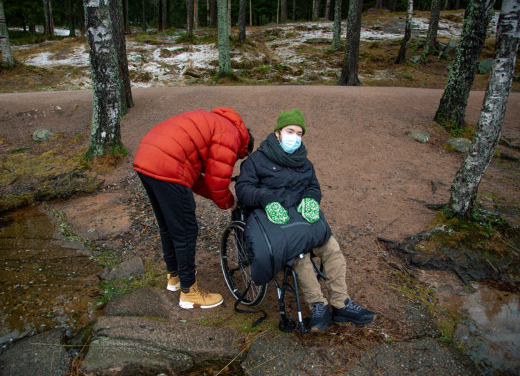 Assistenten til Johannes bøyer seg ned for å fikse noe på rullestolen til Johannes. Rundt dem står det trestammer. Johannes har på seg ansiktsmaske.