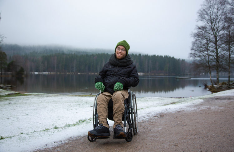 Johannes sitter i en rullestol nede ved vannkanten på Sognsvann. Vi ser hele Johannes. Bak han er bakken hvit og været tåkete.