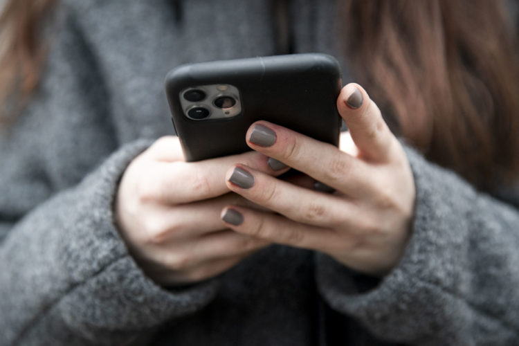 Nærbilde av Synnes hender som holder en iPhone. Vi ser baksiden av telefonen, som har svart deksel. Synne omfavner den, og har på grå- bage neglakk.