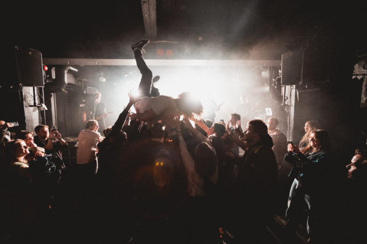 Eit lite konsertlokale er fyllt opp av menneske. Det er mørkt, men lyset frå scena lyser opp siluettane til publikummet. Dei står med armane i været og løfter opp Markus, som akkurat har stagediva frå scenen. 