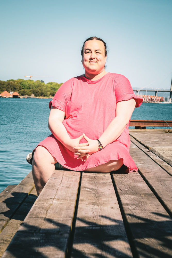 Karoline smiler lurt. Hun sitter på en brygge, med beina i kryss, iført en rosa sommerkjole, og stirrer ut i luften. Været er idyllisk og i bakgrunn ser vi en tydelig blåfarget fjord.