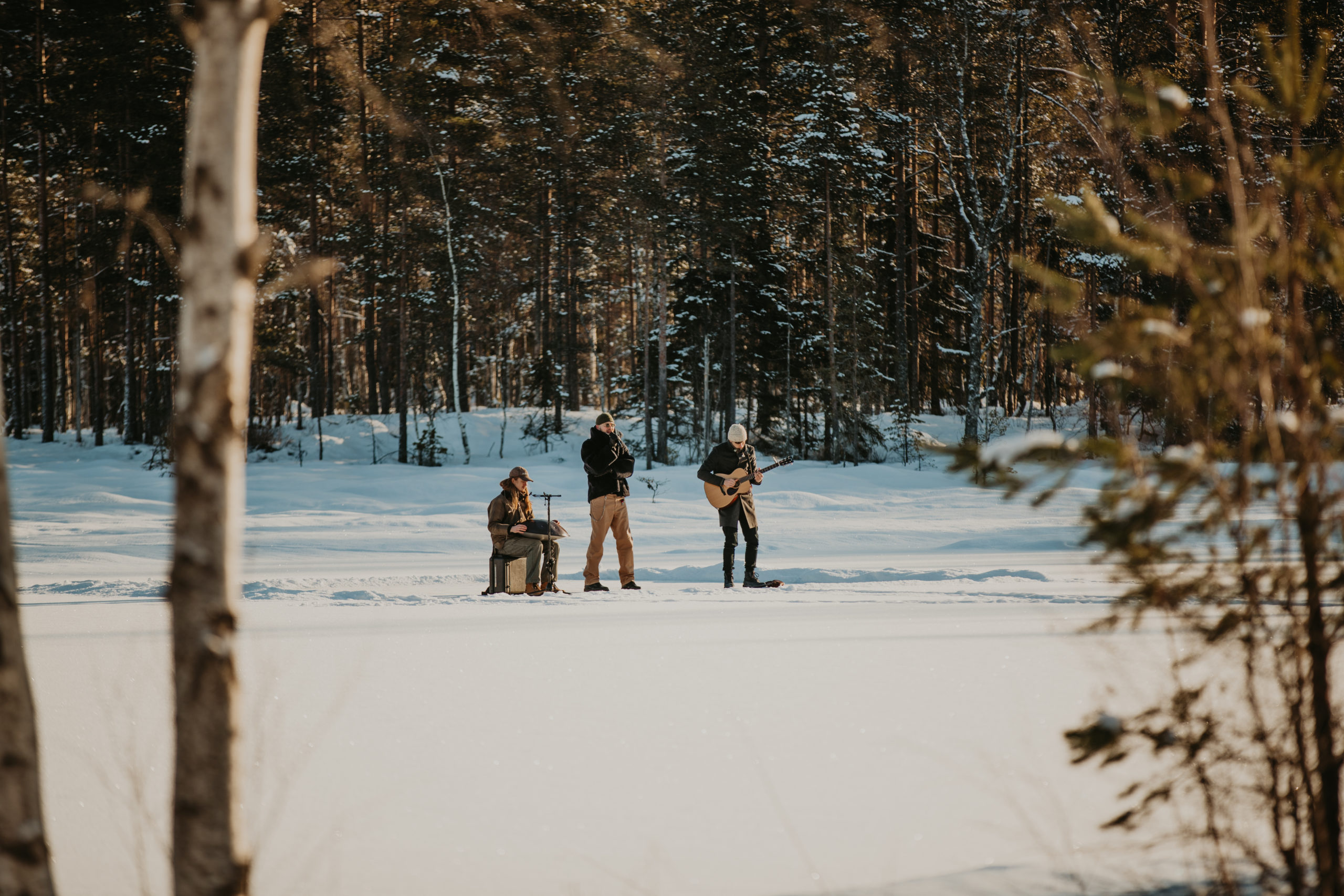 Stig Brenner står sammen med trommis og gitarist på et iskledd vann midt ute i marka. De tre ser små ut der de står langt unna fotografen foran snøkledde trær.