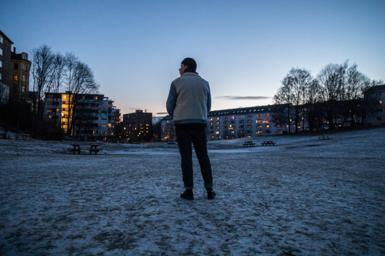 Markus står med ryggen vendt mot kamera i ein park. Det er mørkt rundt han, men det kjem lys frå blokkene framfor han. Det er eit tynt lag med snø på bakken, og himmelen er blå.