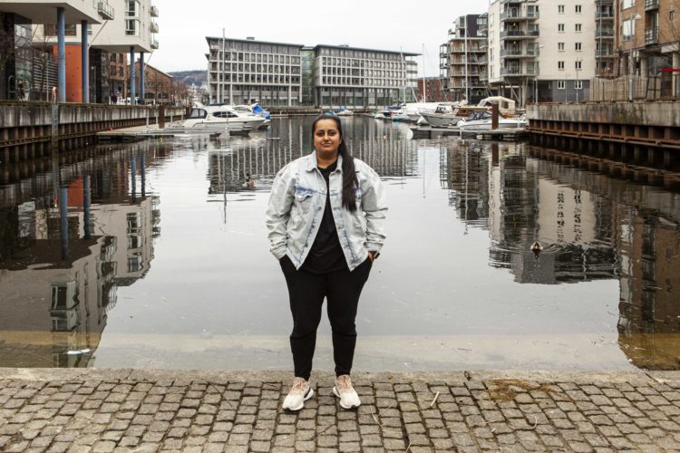 Kiran Anwar står ved vannet på Solsiden i Trondheim. Vi ser henne i helfigur med sorte bukser og dongerijakke på. Hun står med hendene i lomma og er alvorlig i blikket. Bak henne speiler høye bygninger seg i vannflaten. 