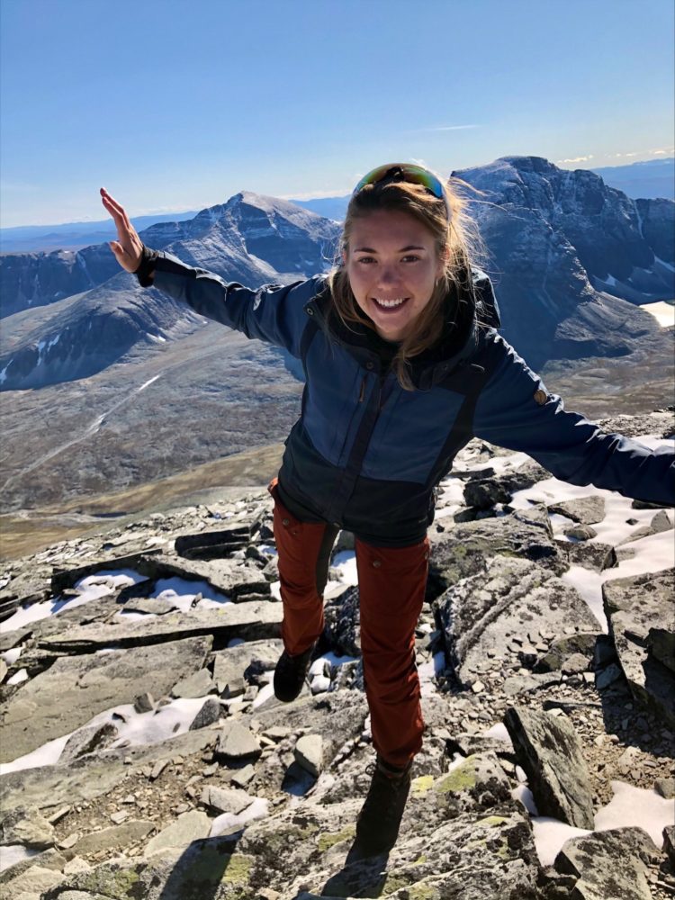 På en ukjent fjelltopp står DNT representanten og balanserer på en fot, med hendene ut i vinger. Bildet er tatt i helprofil og Josephine smiler bredt rett i kameraet.