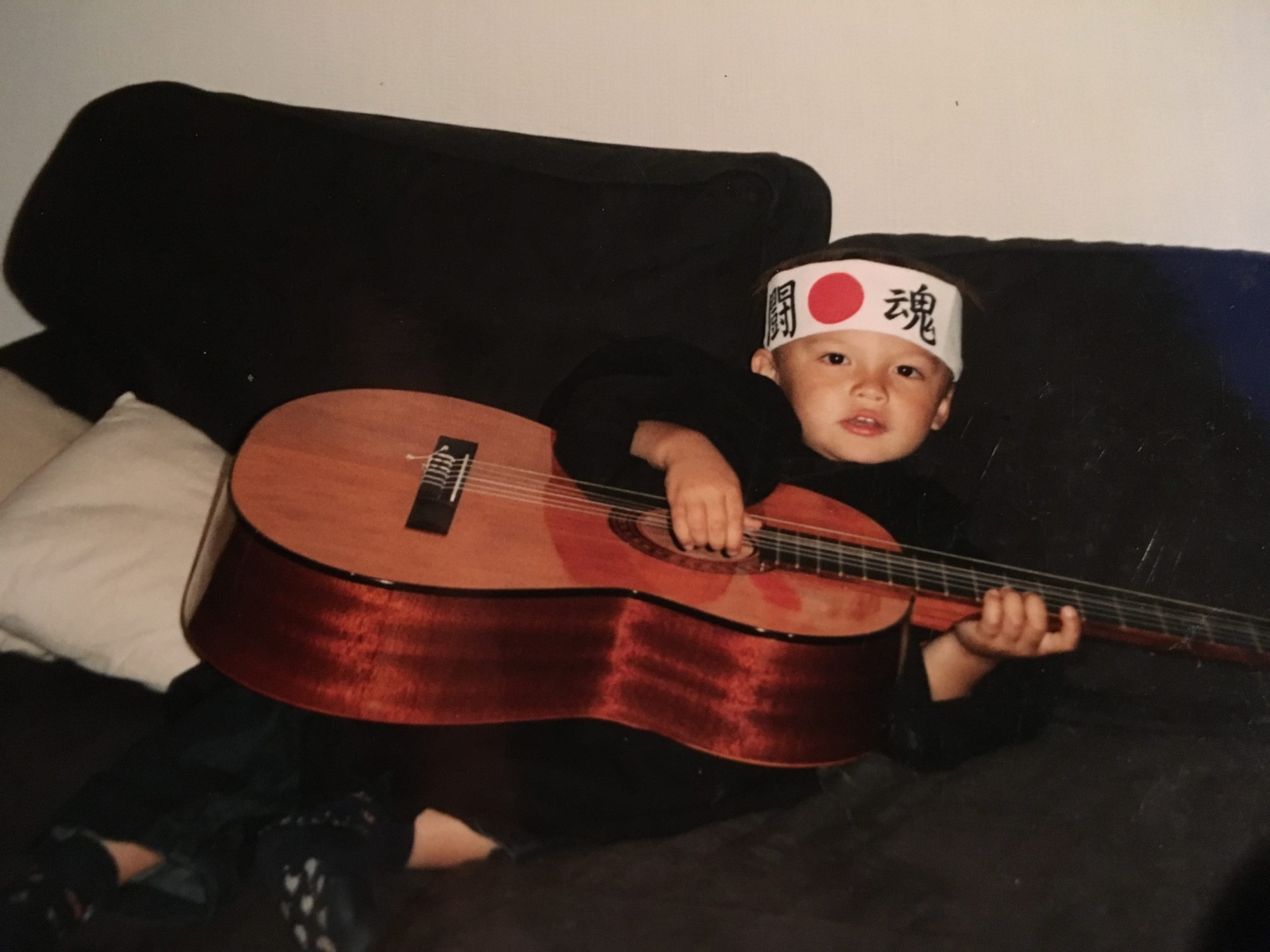 Noah er rundt 5 år gammel. Han ligger i sofaen, med en stor gitar som nesten dekker hele kroppen hans. Han har et panneband med kinesiske tegn på hodet.