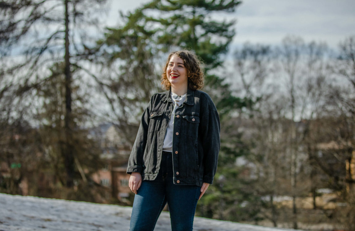 Leona står i ein park. Ho ler og ser ut til sida. Ho har kort, brunt krøllete hår, raud leppestift, blå dongeribukse, grå dongerijakke og ei kvit t-skjorte på seg. Bakken bak ho er dekt av eit tynt lag snø, og bak ho ser ein hus og nokre tre. 