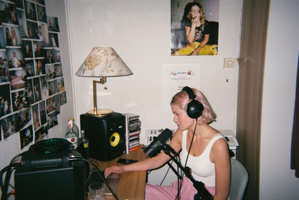 Helene sitter fremfor en mac i et studio. Hun har på seg en hvit topp og rosa bukser. Hun har store hodetelefoner på ørene, en mikrofon fremfor seg og to store høytalere ved siden av macen på pulten. Bak henne står et keyboard og på veggene henger det ulike plakater og bilder. 