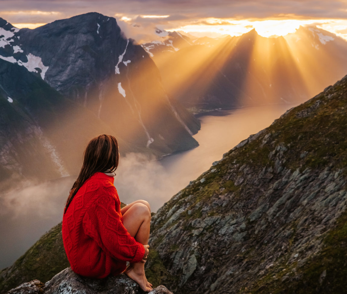 Helene speider ut over et majestetisk fjordlandskap. Solen er på vei ned over de spisse fjellene. Helene sitter med ryggen til kamera på en sten, og ser utover solnedgangen. Håret henger nedover ryggen, og hun har på seg en rød strikket genser. 