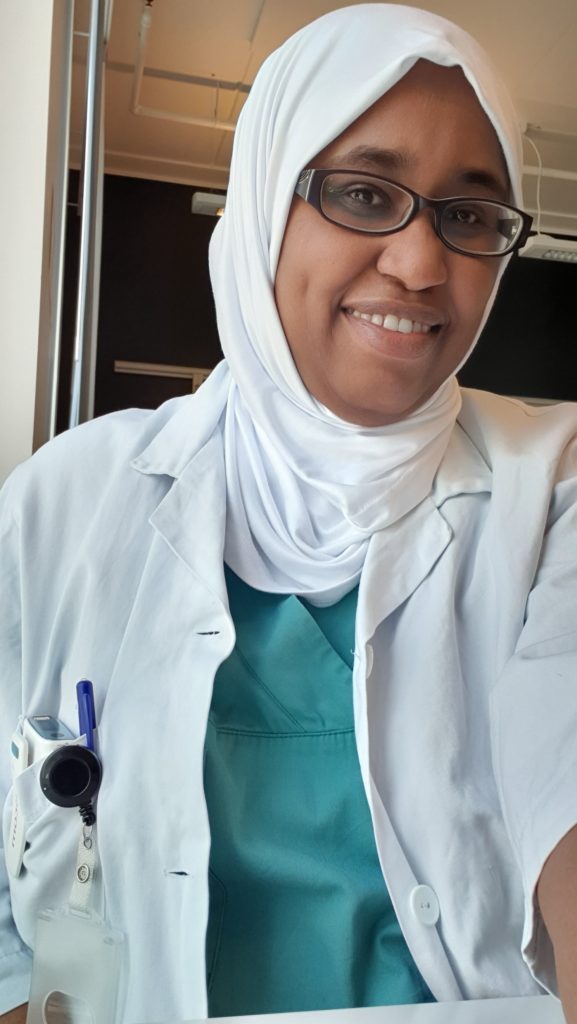 Bilde av lege Samah Abdelkareem som smiler mot kameraet. Hun har legefrakk på med grønne sykehusklær under. Hun har hvit hijab på hodet og briller i ansiktet. I frakken har hun en lomme med en personsøker i og en penn som henger fast i lomma. 
