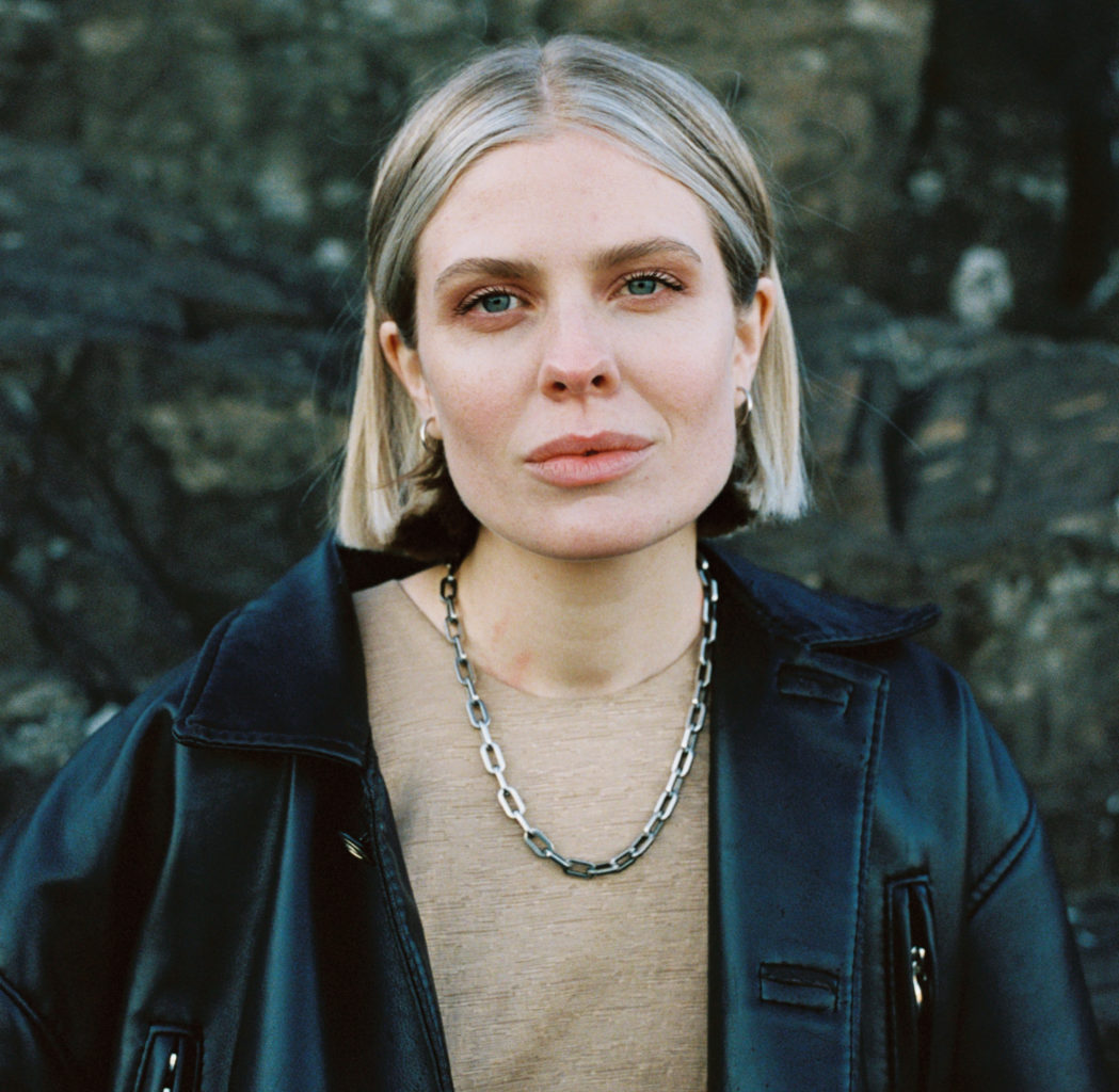 Vokalist i Ponette, Helene Svaland Johansen er kledd i svart jakke og ser i kamera. Ho har kort blondt hår og eit tjukk kjede rundt halsen. 