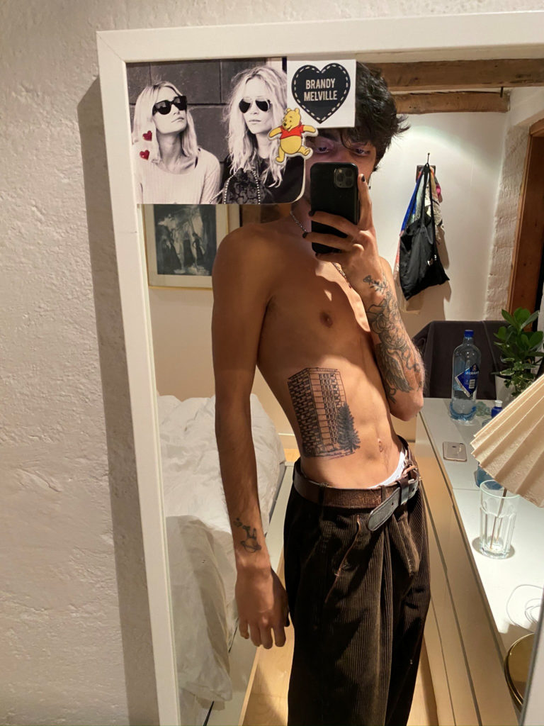 Turab står i bar overkropp og poserer for speilet. Han har en tatovering av en høyblokk på magen, som markerer seg veldig i bildet.