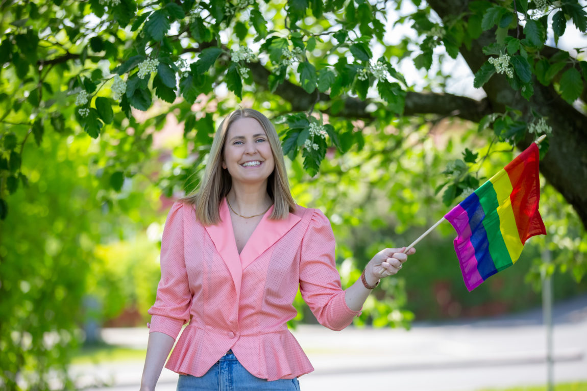 Silje Nordnes ser inn i kamera og smiler. Hun har lyst blondt hår og har på seg en rosa bluse. Hun holder et prideflagg i hånden.